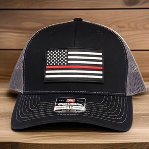 May include: Black and gray trucker hat with a patch featuring a stylized American flag with a red stripe. The hat has a curved brim and a mesh back. The patch reads "Richardson Original 112 Trucker Adjustable".