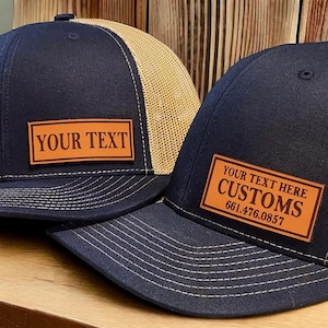 May include: Two black trucker hats with brown leather patches. The patch on the left hat says "YOUR TEXT". The patch on the right hat says "YOUR TEXT HERE CUSTOMS 661.476.0857".