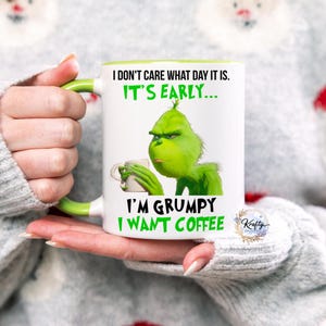 May include: White ceramic mug with a green handle and rim, featuring the Grinch holding a coffee cup. Text on the mug reads "I DON'T CARE WHAT DAY IT IS. IT'S EARLY... I'M GRUMPY I WANT COFFEE."