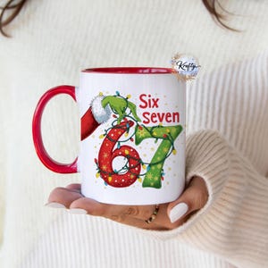 May include: White ceramic mug with a red interior and handle. The mug features the text "Six Seven" with the numbers 6 and 7 decorated with Christmas lights and a Grinch-themed design. A Santa Claus hand is also visible.