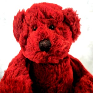 Pode incluir: Um urso de peluche coberto por uma pelagem vermelha vibrante e texturizada. O bicho de peluche apresenta dois olhos pequenos, escuros e redondos, e um nariz preto proeminente e arredondado centrado no rosto. O urso tem orelhas redondas e macias.