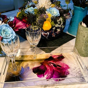 Puede incluir: Una bandeja de servir de acrílico transparente con un diseño floral que presenta una flor rosa. Dos anillos de servilleta dorados con piedras preciosas verdes están sobre una superficie de mármol blanco.