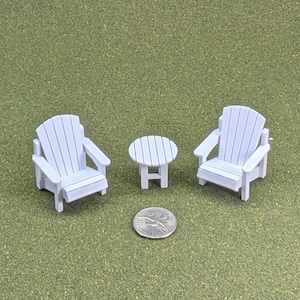 Könnte beinhalten: Zwei weiße Miniatur-Adirondack-Stühle und ein kleiner passender runder Beistelltisch, die zusammen angeordnet sind. Eine silberne Münze ist im vorderen Bereich platziert, um ein klares Gefühl für den kleinen Maßstab dieser Objekte zu vermitteln.