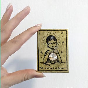 Könnte beinhalten: Kleines, rechteckiges Gemälde mit goldenem Hintergrund und einer schwarzen Illustration einer Frau, die eine Kristallkugel hält. Der Text "THE FUTURE IS BRIGHT" steht unten. Ein kleiner Edelstein befindet sich auf der Stirn der Frau.