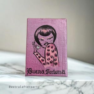 Könnte beinhalten: Kleine, rechteckige Leinwand mit rosa Hintergrund, die eine stilisierte Illustration einer Frau mit schwarzen Haaren und einem Oberteil mit Leopardenmuster zeigt. Der Text "Buena Fortuna" ist in schwarzer Schrift geschrieben. Der Künstlername, @astralafterparty, steht unten.