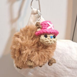 May include: A handmade, fluffy brown plushie with a crocheted face and feet. It wears a pink hat with a white cross and has black eyes and a blue nose. The plushie is attached to a silver keychain.