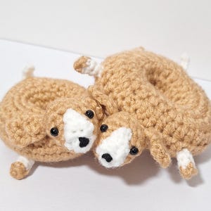 Pode incluir: Dois brinquedos de cão feitos à mão em crochê. Os cães são castanhos com rostos brancos e narizes pretos. Eles têm pequenos olhos pretos e patas e caudas com pontas brancas. Os brinquedos estão em uma superfície branca.