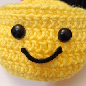 Op de afbeelding: Een geel gehaakt smiley-gezicht met zwarte ogen en een zwarte glimlach.