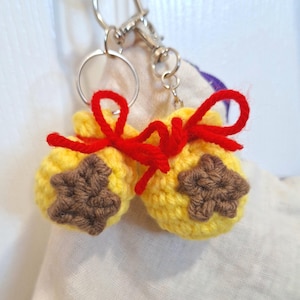 May include: A pair of yellow crocheted pouches with brown star appliqués, tied with red yarn bows, attached to a silver keychain. The pouches are handmade and have a textured appearance, suggesting a craft item.