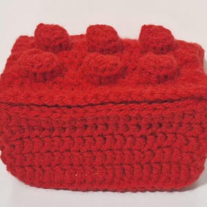 May include: A rectangular red crochet box with a lid featuring six raised circular knobs on top, mimicking the appearance of a building block. The item is constructed from thick red yarn with a visible textured stitch pattern throughout the exterior.