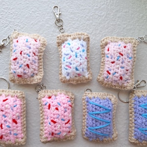 May include: A collection of handmade crochet keychains shaped like Pop-Tarts. The keychains feature pink, white, and purple frosting with colorful sprinkles and blue icing. Each keychain has a metal clasp for easy attachment.