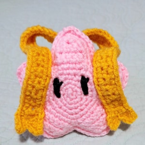 May include: A handmade crochet star-shaped plush toy in pink with black eyes and yellow wings. The toy is made of soft yarn and has a textured appearance. The wings are attached to the back of the star.