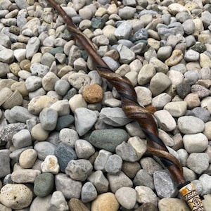 Puede incluir: Una varita de madera hecha a mano con un diseño retorcido, con tonos marrón oscuro y negro. El mango de la varita tiene una punta de color crema con patrones tribales tallados. La varita descansa sobre un lecho de piedras grises y verdes.