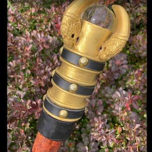 May include: A gold staff with a claw-shaped top grasping a clear orb. The staff features black bands with gold studs and a reddish-brown wooden shaft. The background is filled with green and purple foliage.