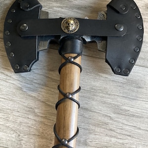 May include: A double-headed axe featuring a patterned Damascus steel blade, a wooden handle wrapped in black leather cord, and black leather blade covers secured with metal rivets. A circular brass-toned skull emblem is centered on the leather strap.