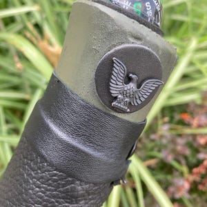 May include: A cylindrical olive green handle featuring a black leather wrap at the base and a circular black emblem with a silver eagle motif. A small round compass with a green directional arrow and white lettering is embedded at the top end.