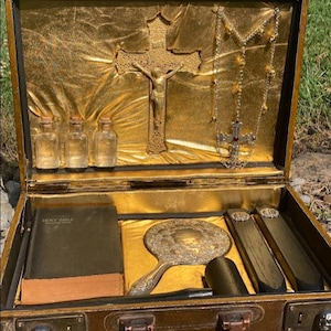 May include: Open brown suitcase with gold interior, holding religious items. Includes a gold crucifix, rosary, small bottles, a bible, a hand mirror, and two rectangular objects. The suitcase has a handle and skull-shaped clasps.