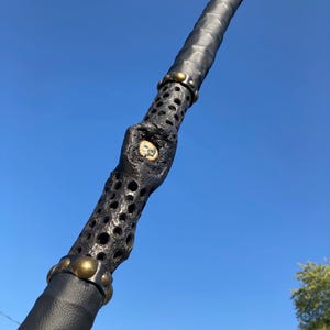 May include: A black leather-wrapped staff with a decorative, hollow, metal section. The metal section has a circular design with gold accents.