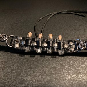 May include: Black leather wristband with four small glass vials, each sealed with a cork stopper. The band features silver-toned metal studs and decorative skull accents on each end. A dark, gothic-inspired accessory.