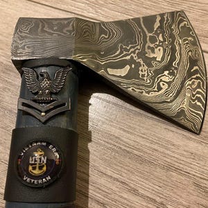 May include: A black and silver axe with a patterned blade. The handle is wrapped in black leather with a US Navy veteran patch and an eagle insignia.