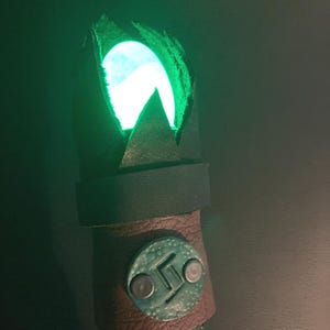 May include: A handcrafted staff with a glowing green orb at the top. The staff has a dark leather band with a decorative metal emblem and a textured wooden handle. The orb emits a bright green light, creating a mystical effect.
