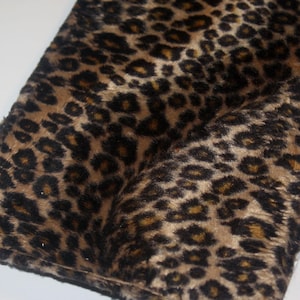 May include: A close-up shot of a folded leopard print fabric. The fabric has a tan base with dark brown and black spots. The material looks soft and plush, with a slight sheen.
