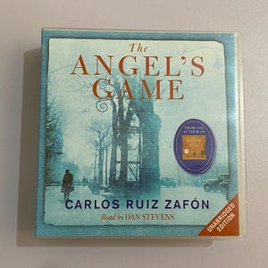May include: A book titled "The Angel's Game" by Carlos Ruiz Zafón, read by Dan Stevens. The cover features a muted blue and gray illustration of a city scene with text in orange. Includes a sticker that says "From the author of Shadow of the Wind".