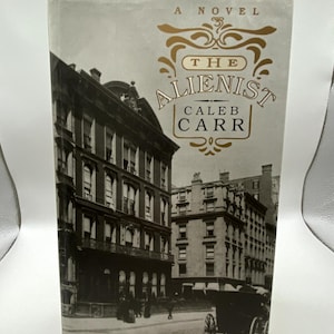 Op de afbeelding: Een hardcover boek getiteld "The Alienist: A Novel" van Caleb Carr. De omslag toont een zwart-wit afbeelding van een stadsstraat met gebouwen en een paardenkoets. De titel staat in gouden letters.