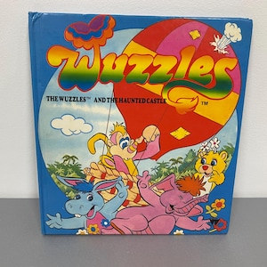Könnte beinhalten: Ein Vintage-Kinderbuch mit dem Titel "The Wuzzles and the Haunted Castle". Das Cover zeigt bunte Zeichentrickfiguren, einen Heißluftballon und den Titel in großen, mehrfarbigen Buchstaben auf blauem Hintergrund.