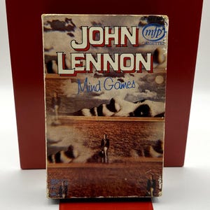 Puede incluir: Una caja de casete rectangular para el álbum Mind Games de John Lennon. La portada presenta un paisaje surrealista con una figura de pie en un campo bajo un cielo nublado. El texto dice JOHN LENNON, Mind Games e incluye el logotipo de mfp.