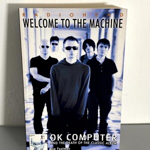 May include: A book titled "Welcome to the Machine" by Radiohead. The cover features a group of five men in dark clothing and sunglasses. The book's title is in large white letters, with the subtitle "OK Computer and the Death of the Classic Album" and the author's name, Tim Footman.