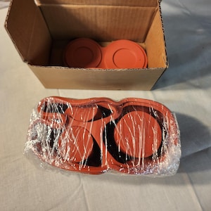 May include: An open cardboard box containing two orange circular objects. Below the box, a package wrapped in clear plastic contains several more orange circular objects. The objects appear to be automotive parts.