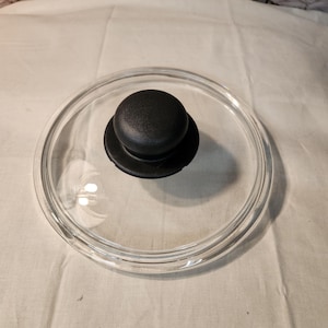 May include: A circular, transparent glass lid for a pot or pan, featuring a smooth, rounded black knob handle centered on top. The lid has a clear glass surface with a slightly raised outer rim, designed to fit securely onto cookware.