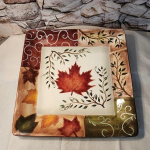 May include: A square ceramic platter featuring a central red maple leaf on a cream background, surrounded by a border of autumn-themed leaves in shades of brown, yellow, and green, accented with delicate vine patterns and swirling white lines.