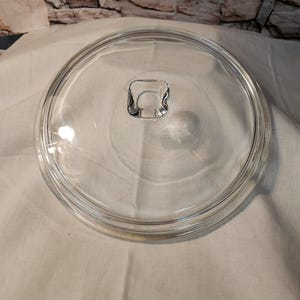 May include: A clear glass lid with a rounded shape and a small, square handle in the center. The lid is designed to fit on a pot or pan. The background is a light-colored fabric.