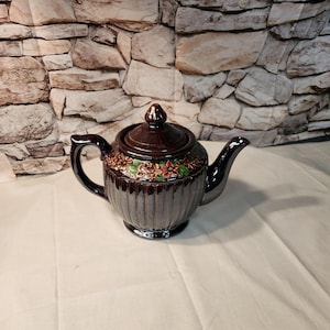 May include: A dark brown ceramic teapot with a glossy finish and a vertical ribbed body. The upper section features a decorative band of embossed flowers in shades of orange, green, and brown. It includes a matching lid with a small knob handle.