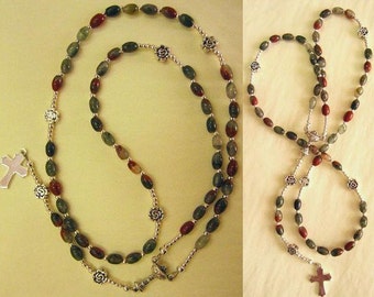 May include: A rosary with oval-shaped beads in shades of green, red, and brown. The beads are strung on a silver-colored chain, with silver cross and floral accents. The rosary is displayed against a light yellow background.