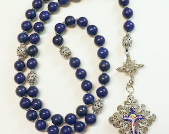 May include: A rosary featuring deep blue spherical beads interspersed with ornate silver-toned filigree metal spacers. The piece concludes with a detailed silver-toned filigree cross containing a central blue enamel inlay depicting a figure.