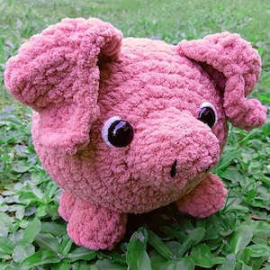 May include: A handmade, plush pig in a soft pink hue. The pig has large, black eyes, a small snout, and floppy ears. It is sitting on a bed of green foliage, creating a charming, whimsical scene.