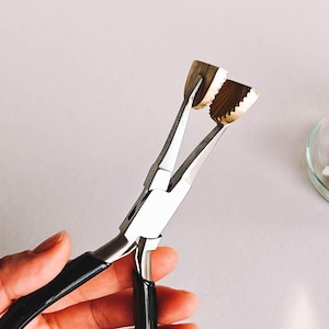 May include: Jewelry pliers with black handles and silver metal jaws. The jaws have a textured surface. The pliers are held in a hand against a white background. A glass cloche with decorative items is in the background.