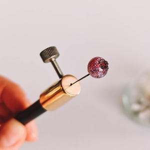Könnte beinhalten: Nahaufnahme eines Schmuckwerkzeugs, das eine kleine, runde, rote Perle mit schwarzen Akzenten hält. Das Werkzeug hat einen schwarzen Griff, eine goldfarbene Metallbasis und eine silberne Schraube. Der Hintergrund ist eine weiche, neutrale Farbe.