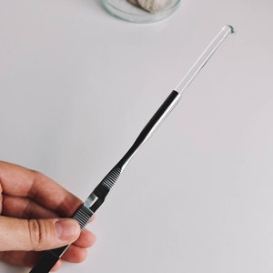 May include: A silver-toned, medical-style tweezer with a clear, rounded tip. The tweezer has a ridged grip and a sleek, modern design. The tool is held against a white background.