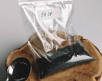 May include: A clear plastic bag with a label that says "54 SP" contains a dark gray powder. The bag is sitting on a wooden surface.