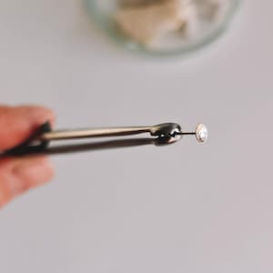 Puede incluir: Una pequeña gema redonda y transparente sostenida por unas pinzas plateadas. Las pinzas están enfocadas, con la gema centrada. El fondo es de un color suave y neutro, con un recipiente de vidrio borroso visible.