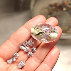 Op de afbeelding: Een grote ovale cabochon van helder glas met een ingebed bloemmotief met paarse bloemblaadjes en groene stengels, vergezeld door verschillende kleinere, ronde, klokvormige glaskralen in wit en paars met gestreepte patronen.