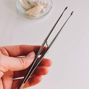Pu&ograve; includere: Pinzette in acciaio inossidabile con manici strutturati e punte affilate. Le pinzette sono tenute in una mano, con un piccolo piatto di vetro contenente una sostanza di colore chiaro sullo sfondo. Per lavori di precisione.
