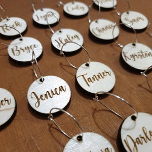 Puede incluir: Un juego de 12 charms para copa de vino personalizados con nombres grabados en etiquetas redondas de madera. Los charms están unidos a aros de metal plateado.