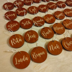 Puede incluir: Un conjunto de 20 charms redondos de madera para copas de vino con nombres escritos en letras doradas. Los charms están unidos a anillos plateados.