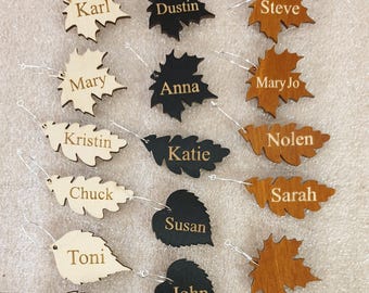 Puede incluir: Un conjunto de 15 porta tarjetas de mesa de madera en forma de hoja con nombres grabados. Las hojas son de varios tonos de marrón, negro y madera natural. Los nombres incluyen Karl, Dustin, Steve, Mary, Anna, Mary Jo, Kristin, Nolen, Katie, Chuck, Sarah, Susan, Toni, John y Rick.