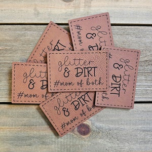May include: A set of five brown leather tags with the text "glitter & dirt #mom of both" printed in black. The tags have a stitched border.